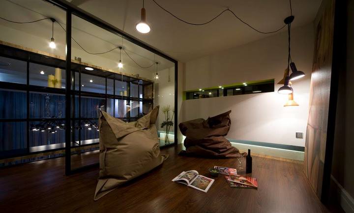 This stylish lounge area by Design Spirits showcases a chic industrial theme from their Commercial: SOHO Empire portfolio. It features a warm, dark wood floor contrasted with minimalist furniture and strategic lighting. The space is defined by glass partitions with black metal frames, complementing the suspended Edison bulbs above, evoking a modern, yet cozy ambiance perfect for both relaxation and social gatherings, exemplifying contemporary Malaysian interior design.