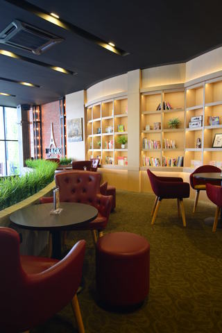 Contemporary meets rustic in this cozy cafe corner by Young Concept Design Sdn Bhd, showcasing a harmonious blend of warm earth tones and rich textures at Black Tower Coffee in Cheras. Elegant button-tufted armchairs in a deep cherry hue invite patrons to relax, while the subtle carpet pattern complements the bespoke wooden bookshelves. Natural light filters through vertical blinds, casting a soft glow on the curated selection of books and decorative items, epitomizing the detailed interior design and carpentry works.