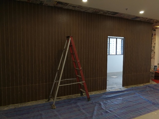 An office space in the midst of a chic renovation by Funlink Creative Sdn Bhd, showcasing a modern design with a deep brown textured wall, complemented by clean lines and natural light peeking through a side window. The silver ladder and protective floor coverings hint at the transformation in progress, representing the meticulous attention to detail characteristic of Funlink's portfolio.