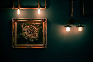 This image captures the essence of the 'HUGO STEAMPUNK PREMIUM LOUNGE' by BANGKU STORE, exemplifying Malaysia's premier steampunk-themed bar. The interior showcases a pseudo-Victorian backdrop with a dark teal wall, adorned by a classic framed artwork depicting intricate mechanical gears, complementing the industrial design theme. Dual exposed bulb fixtures with brass fittings enhance the retro-futuristic atmosphere, perfect for design enthusiasts seeking a fusion of historical elegance and imaginative technofantasy.