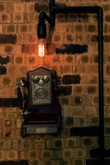 This image showcases a corner of the HUGO STEAMPUNK PREMIUM LOUNGE by BANGKU STORE, highlighting a vintage-inspired phone and exposed Edison bulb against a rustic brick wall. The design captures the essence of a steampunk theme with its dark, moody tones, and mechanical details, embodying BANGKU STORE's luxurious interpretation of the classical railway station aesthetic for the sophisticated interior design enthusiast.