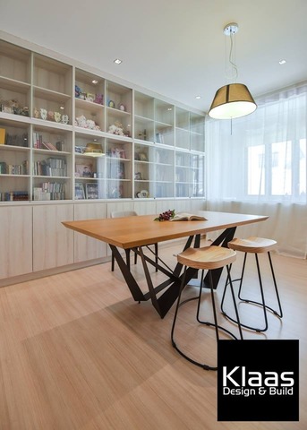 A modern home office by Klaasmen Sdn Bhd in a Bandar Puteri terrace renovation showcases a sleek, minimalistic design. The room features light wood floors, a spacious white shelving system, and a large, natural wood desk paired with black stools. The contemporary space is complemented by a stylish pendant light and sheer white curtains, creating a bright and airy atmosphere, perfect for productivity and creativity within a sophisticated interior.